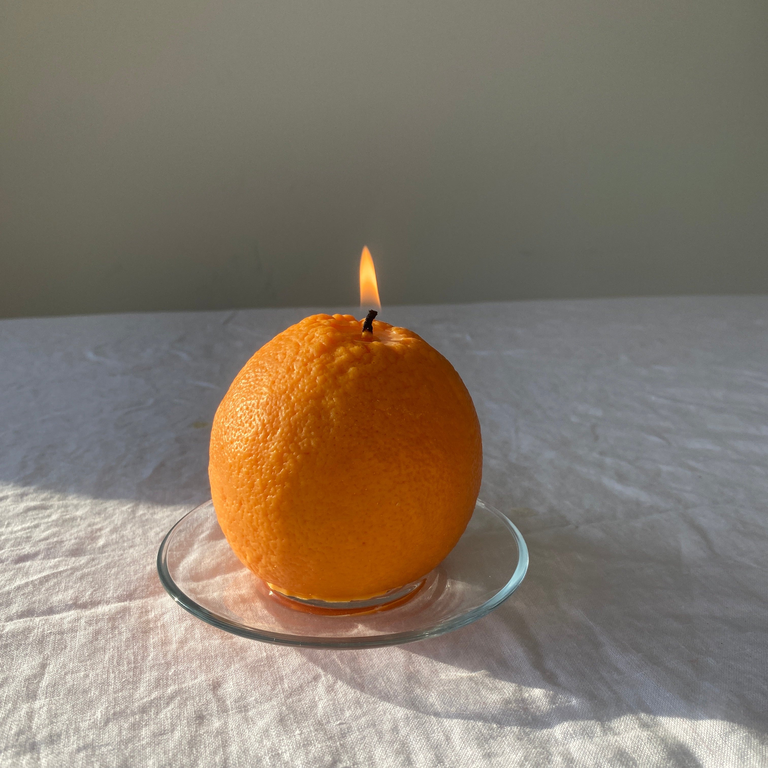 An orange-shaped candle with a visible flame sits on a small clear glass plate on a white fabric surface against a plain light gray wall.