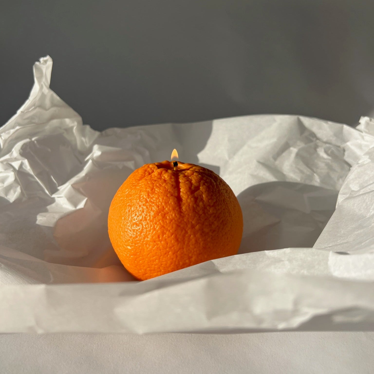 A single orange candle with a small visible wick sits on crumpled white paper against a dark gray background.