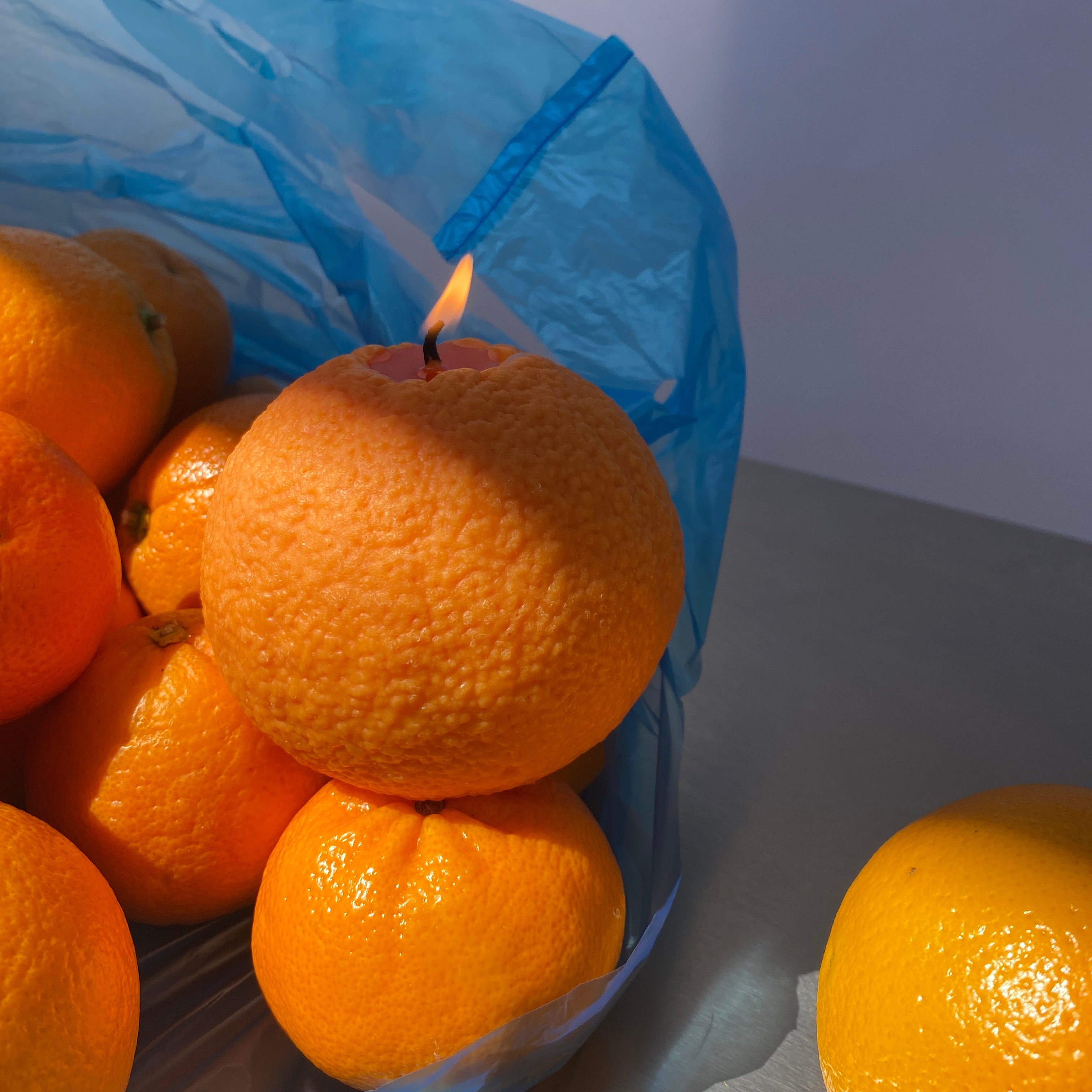 Several bright oranges in a blue plastic bag on a light gray surface, with one orange partially visible at the top right.
