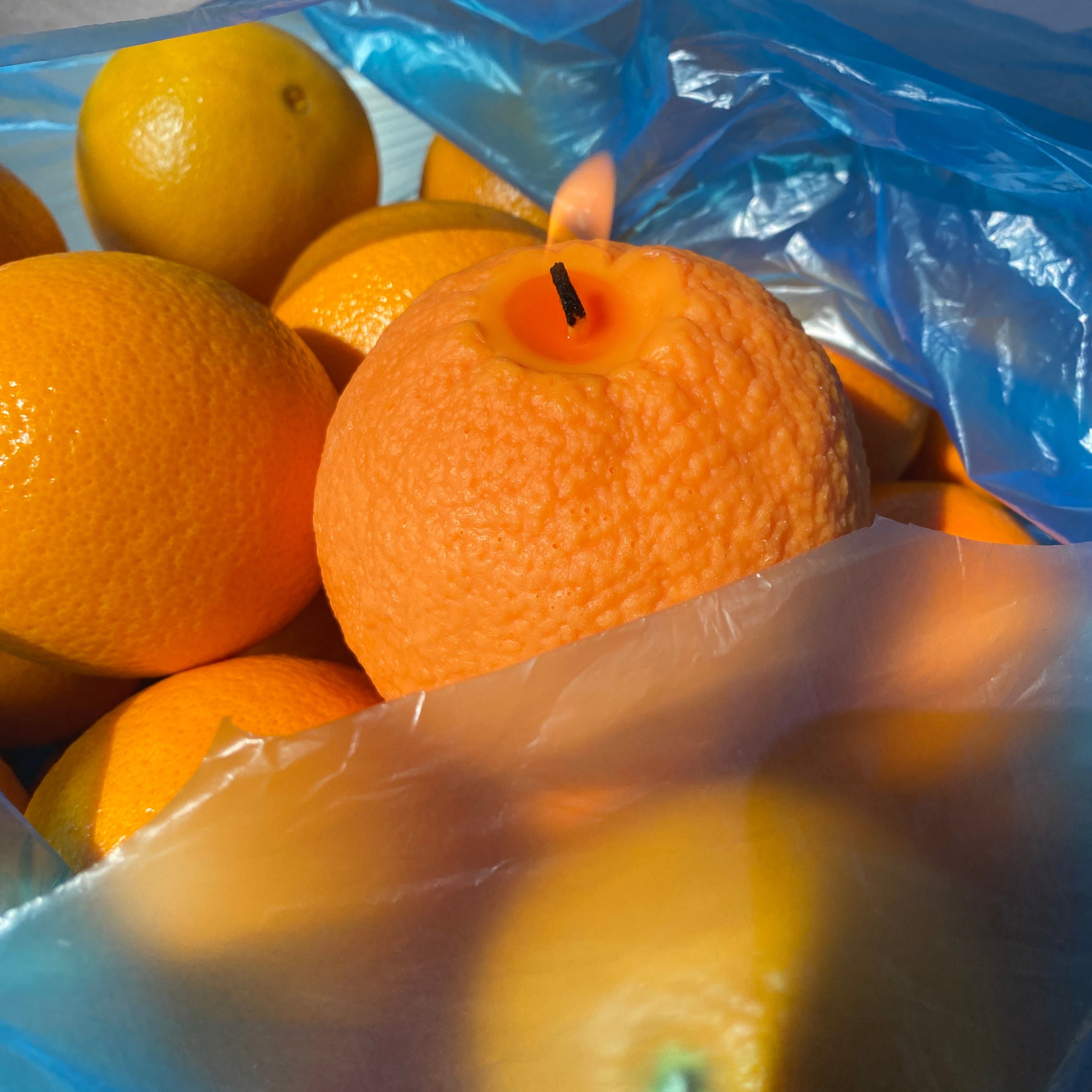 A lit orange candle surrounded by whole oranges inside a blue plastic bag.